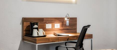 A modern hotel room with a wooden desk, a black chair, a coffee maker, and a flat-screen TV mounted above the desk.