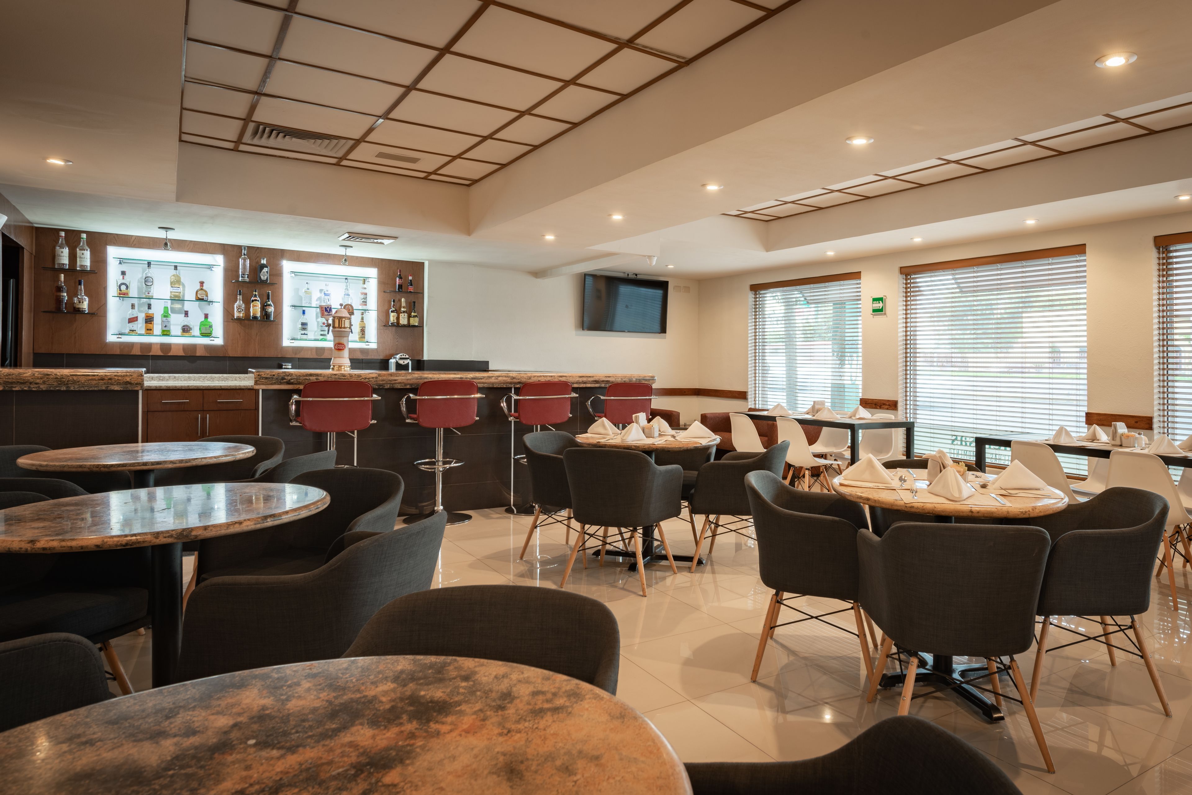 A modern restaurant with round tables and chairs, a bar area with bottles, and a TV mounted on the wall.