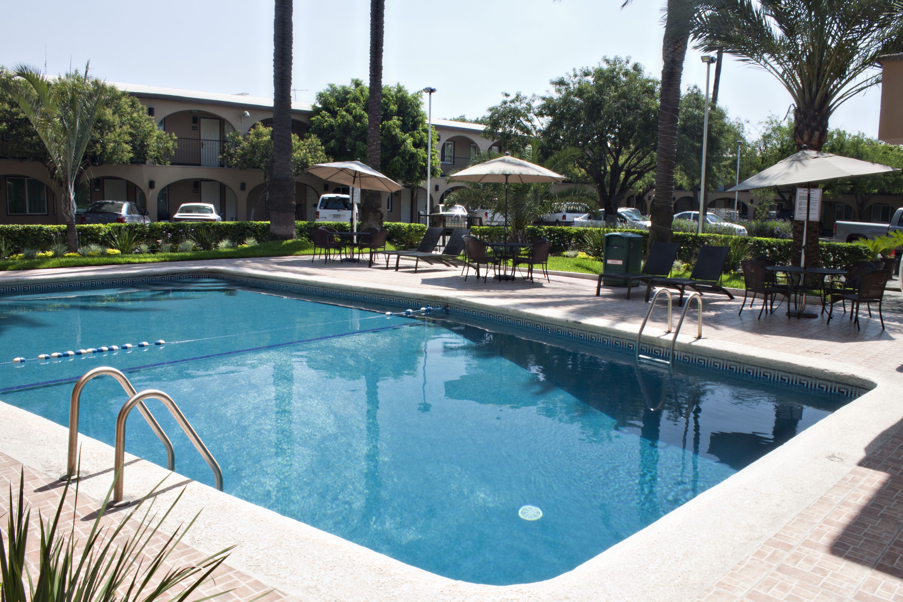 Una piscina con sillas de descanso y sombrillas, rodeada de palmeras y un edificio con balcones.