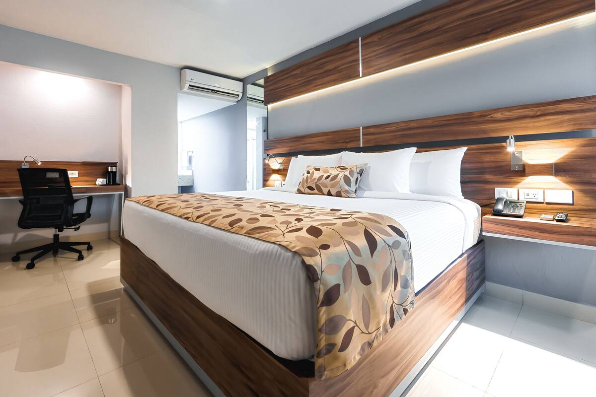 Una habitación de hotel moderna con una cama grande, un escritorio con una silla, una mesita de noche con una lámpara y un equipo de aire acondicionado.