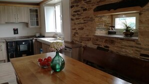 Une cuisine aux murs de pierre, une table en bois ornée d’un vase et de tomates, et une horloge au mur.