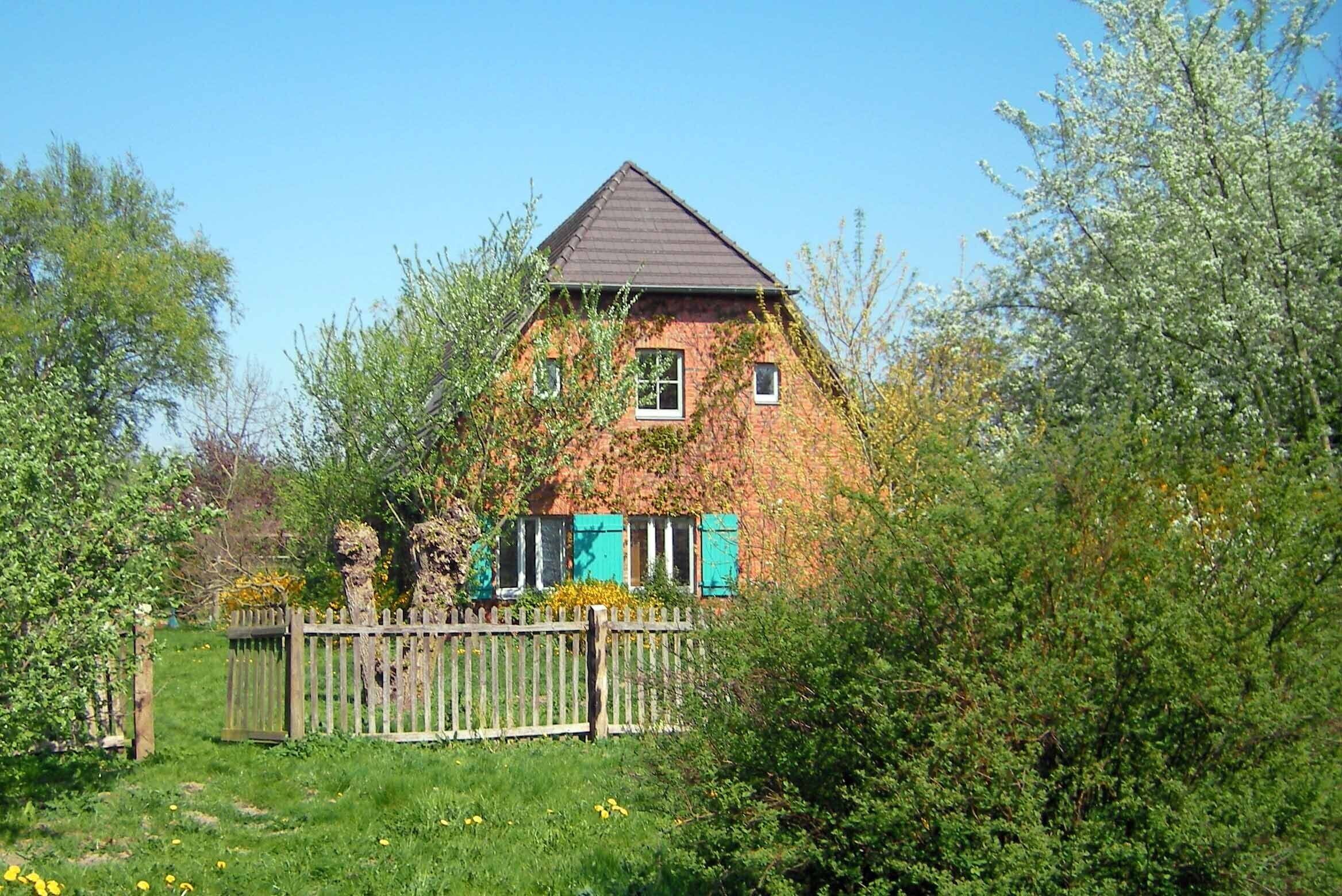 A brick house with a gabled roof, surrounded by greenery and a wooden fence.