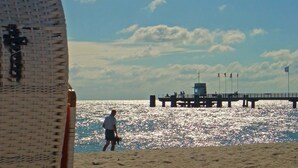 A beach with a pier extending into the water, a man walking on the sand, and a large woven structure in the foreground. - Spacious holiday home with a large garden and swimming pond (Stepenitztal)