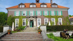 A large, two-story building with a brick facade, green shutters, and a red-tiled roof. - Spacious holiday home with a large garden and swimming pond (Stepenitztal)