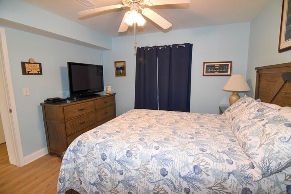 A bedroom with a bed, a television on a wooden dresser, a ceiling fan, and two framed pictures on the wall.