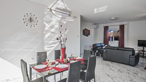 A modern dining area with a glass table, red placemats, and a chandelier. There is a gray sofa and a television in the background. - Your Dream Holiday Villa with Private Pool in Protaras’s most Exclusive Neighbourhood (Protaras)