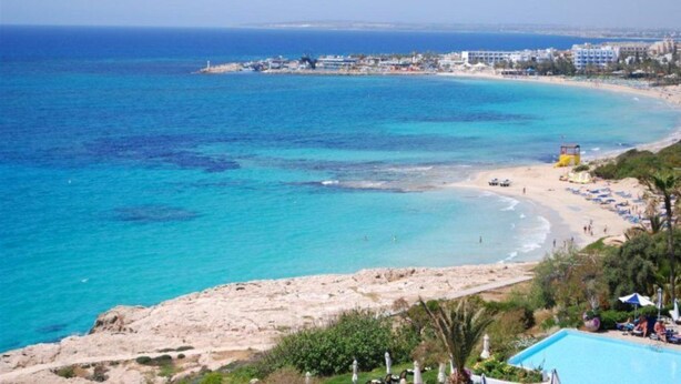 A coastal view with a sandy beach, a clear blue sea, and a resort area with buildings and a swimming pool. - Your Dream Holiday Villa with Private Pool in Protaras’s most Exclusive Neighbourhood (Protaras)