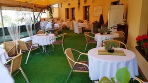 Un área de comedor al aire libre con mesas y sillas redondas, cubierta por un toldo amarillo y piso de césped artificial verde.