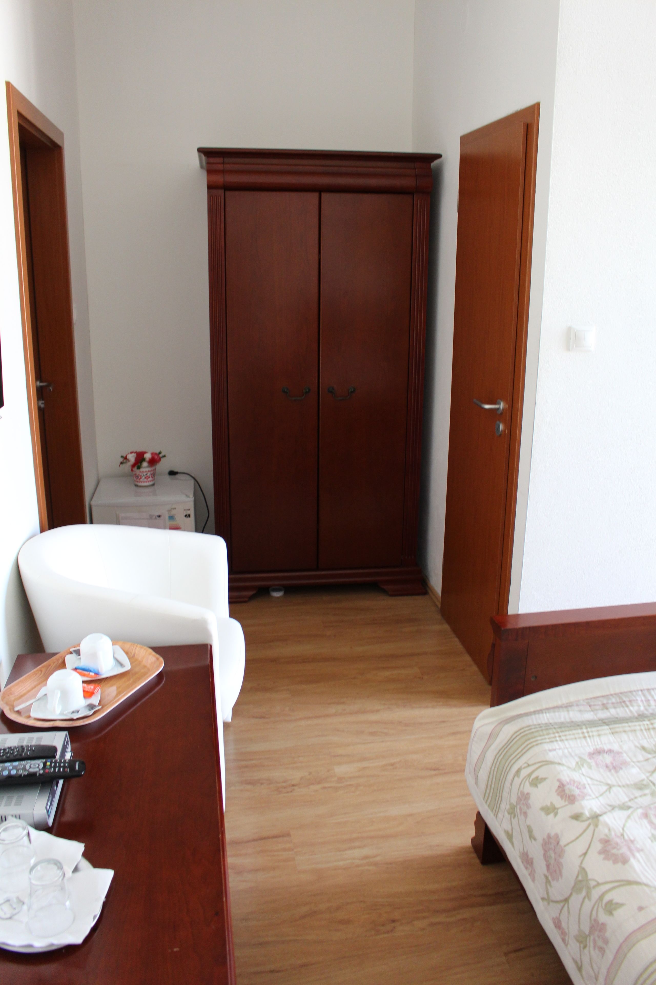 A hotel room with a wooden wardrobe, a white armchair, a bed with floral bedding, and a small table with a tray of glasses and a remote control.