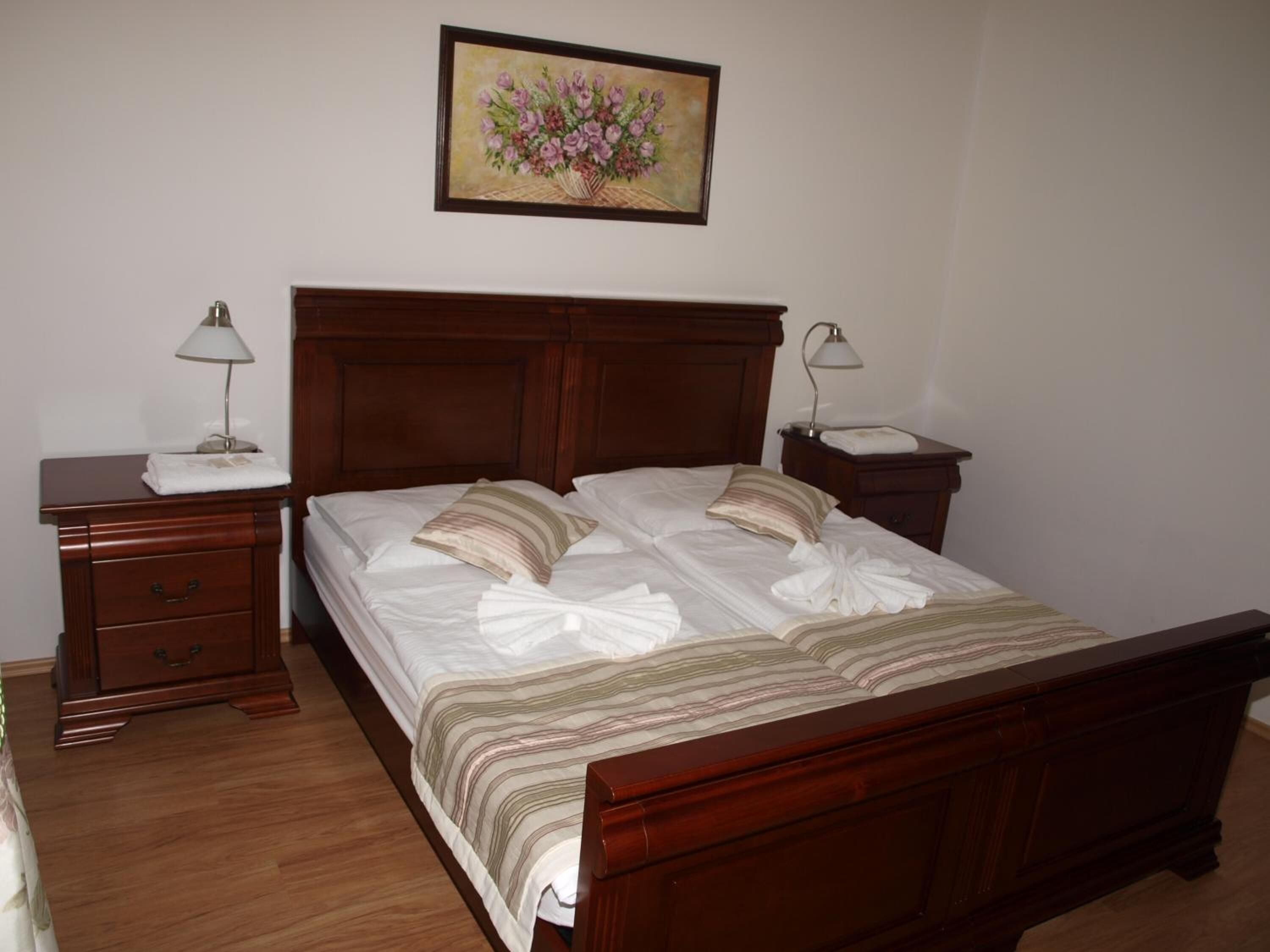 A neatly made bed with a striped bedspread and two pillows, a wooden nightstand with a lamp, a wooden dresser, and a framed floral painting on the wall.