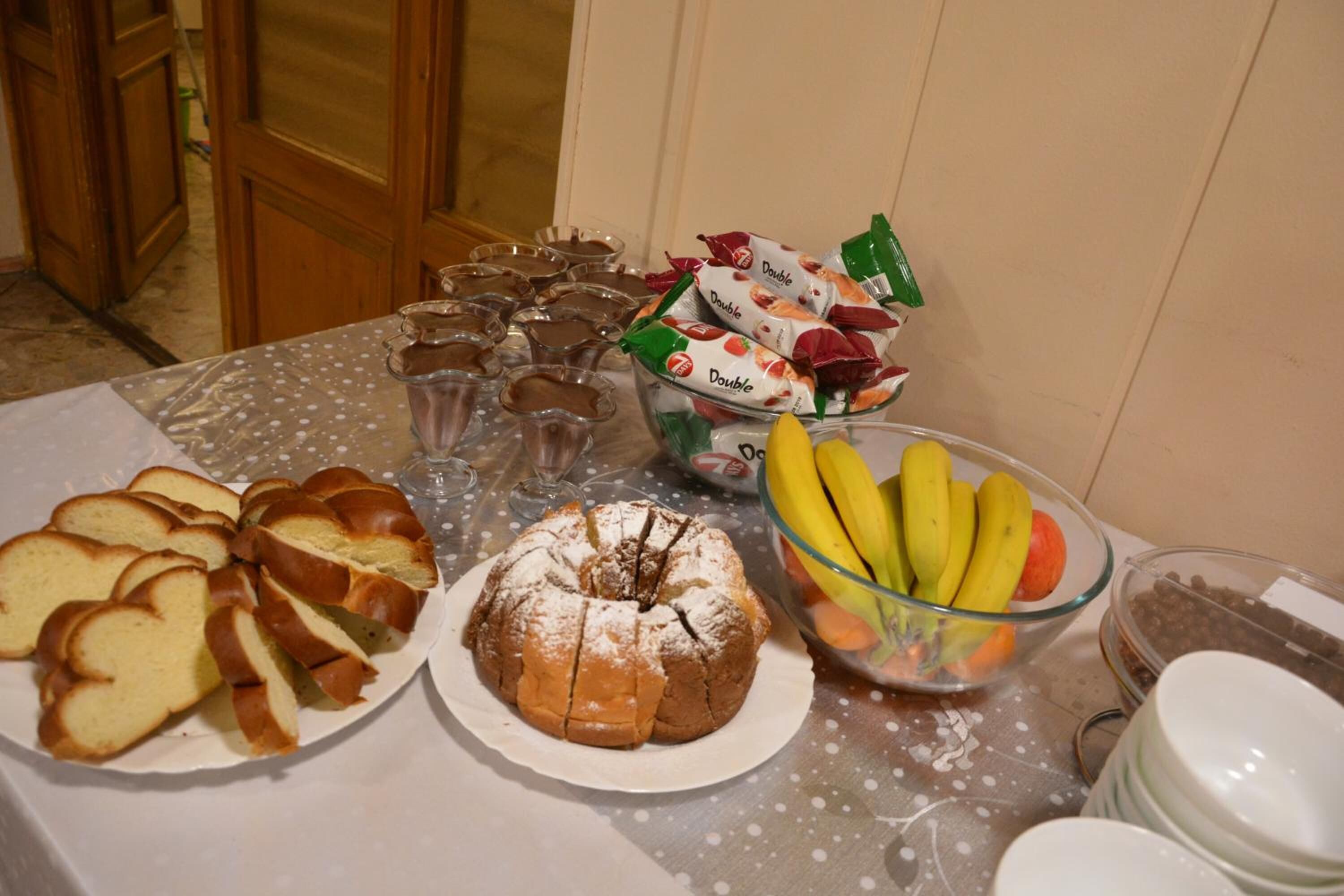 Una mesa con diversos alimentos, como pan en rebanadas, un pastel bundt, un cuenco de fruta y snacks empaquetados.
