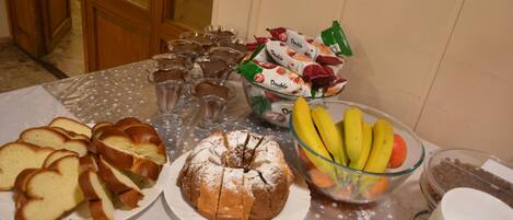 Una mesa con diversos alimentos, como pan en rebanadas, un pastel bundt, un cuenco de fruta y snacks empaquetados.