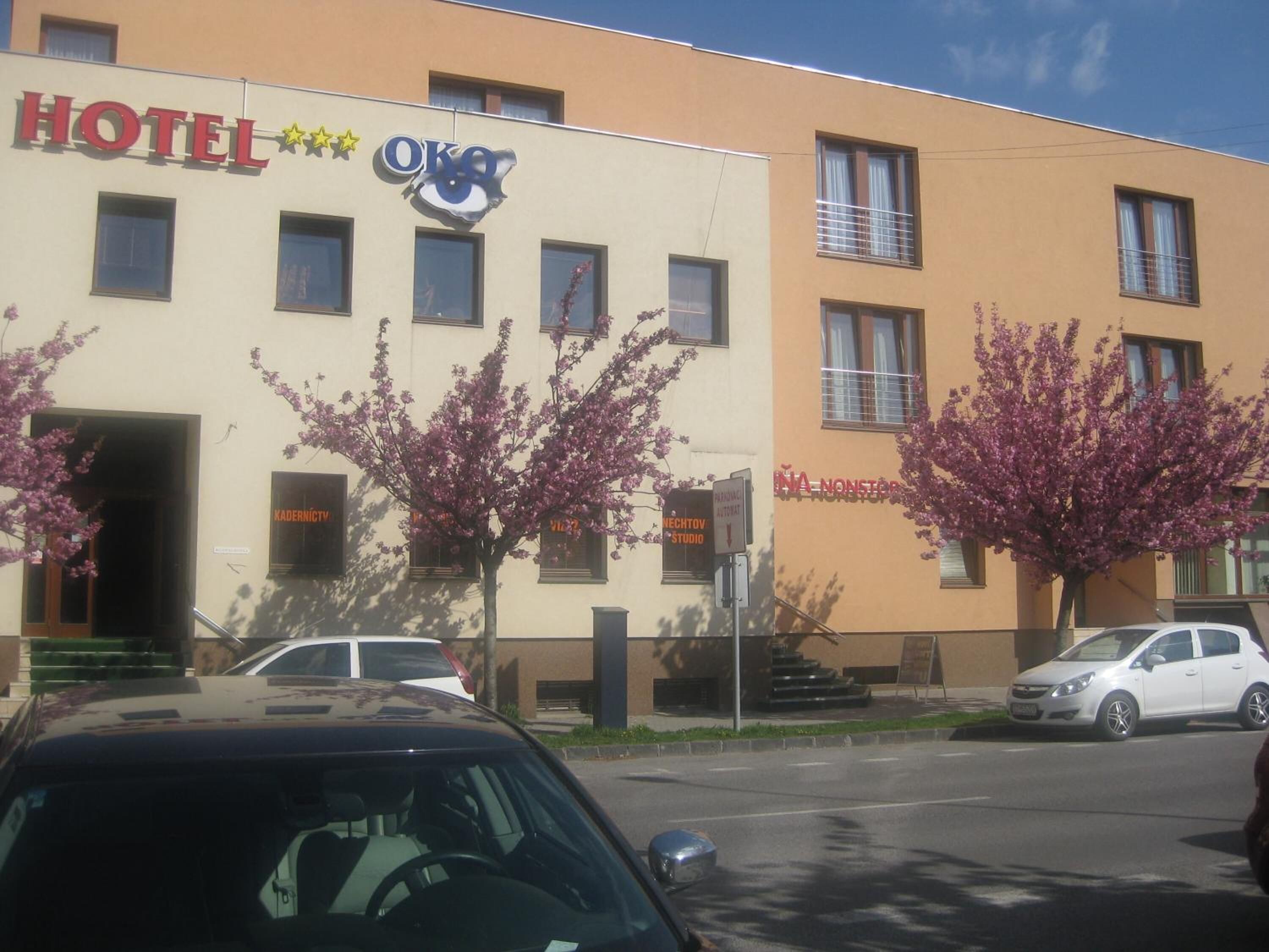 A hotel building with a sign that reads "HOTEL OKO". There are parked cars in front of the building.