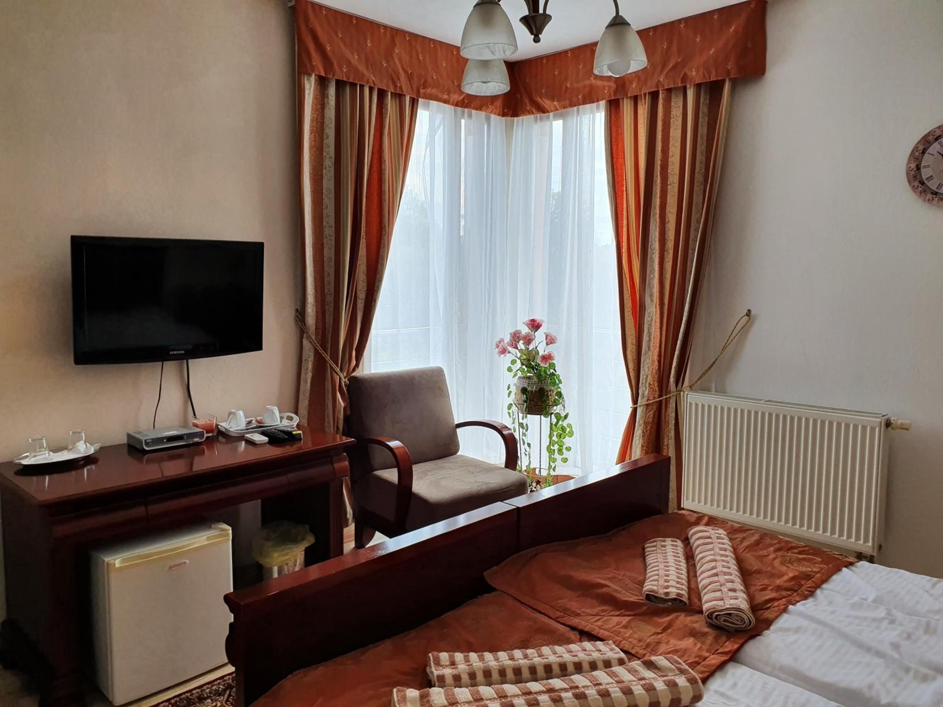 A hotel room with a bed, a desk with a television, a chair, a small refrigerator, and a radiator.