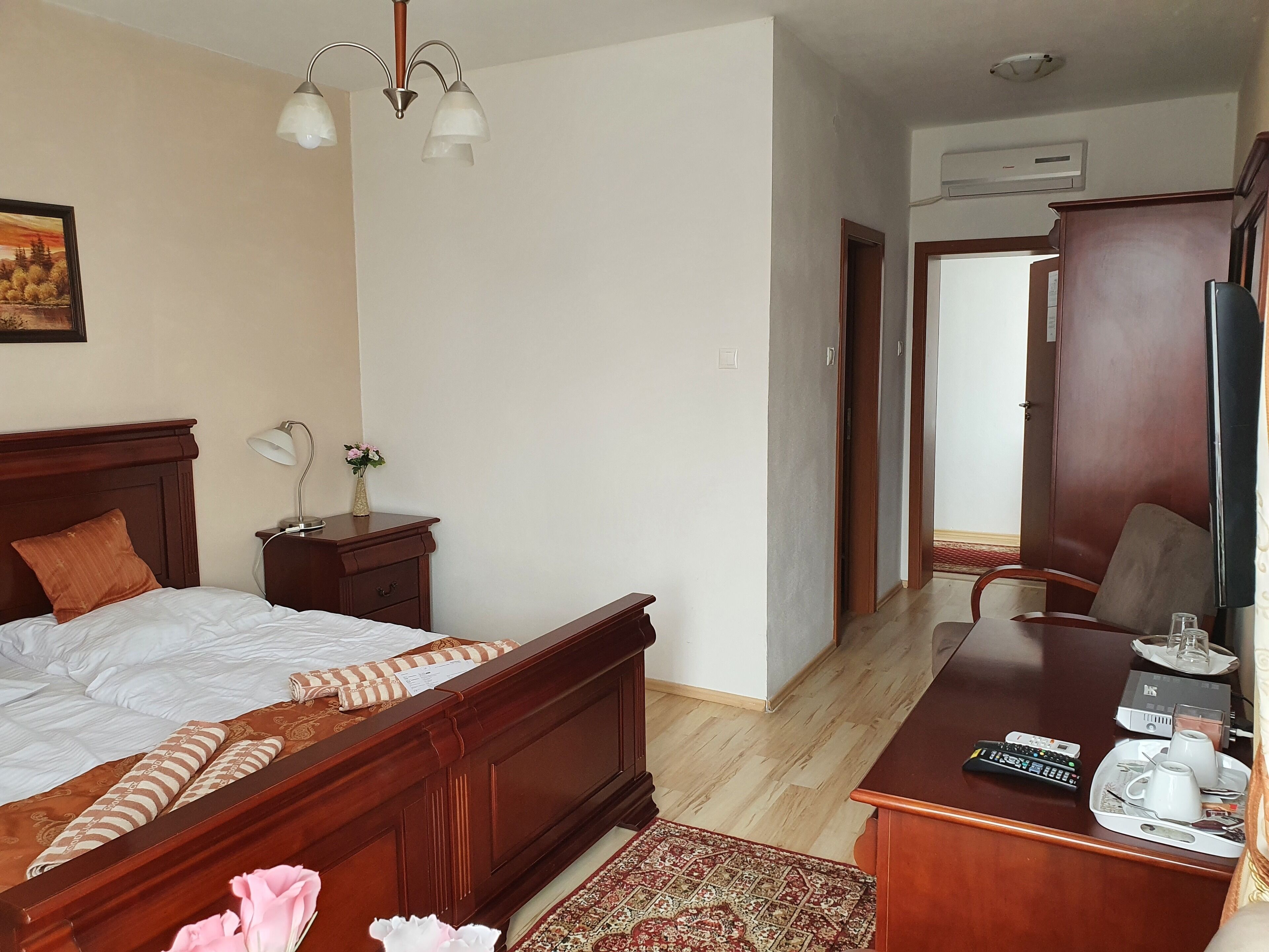 A hotel room with a wooden bed, bedside table, lamp, and a desk with a TV, remote controls, and a tray with cups and a glass.