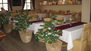 Een eetgedeelte met gedekte tafels voor gasten, met geweven manden met planten, witte tafelkleden en een bar met flessen op de achtergrond.