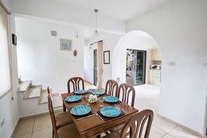 A dining area with a wooden table set for four, surrounded by chairs. There is a staircase leading to an upper level, and a kitchen area is visible through an archway.