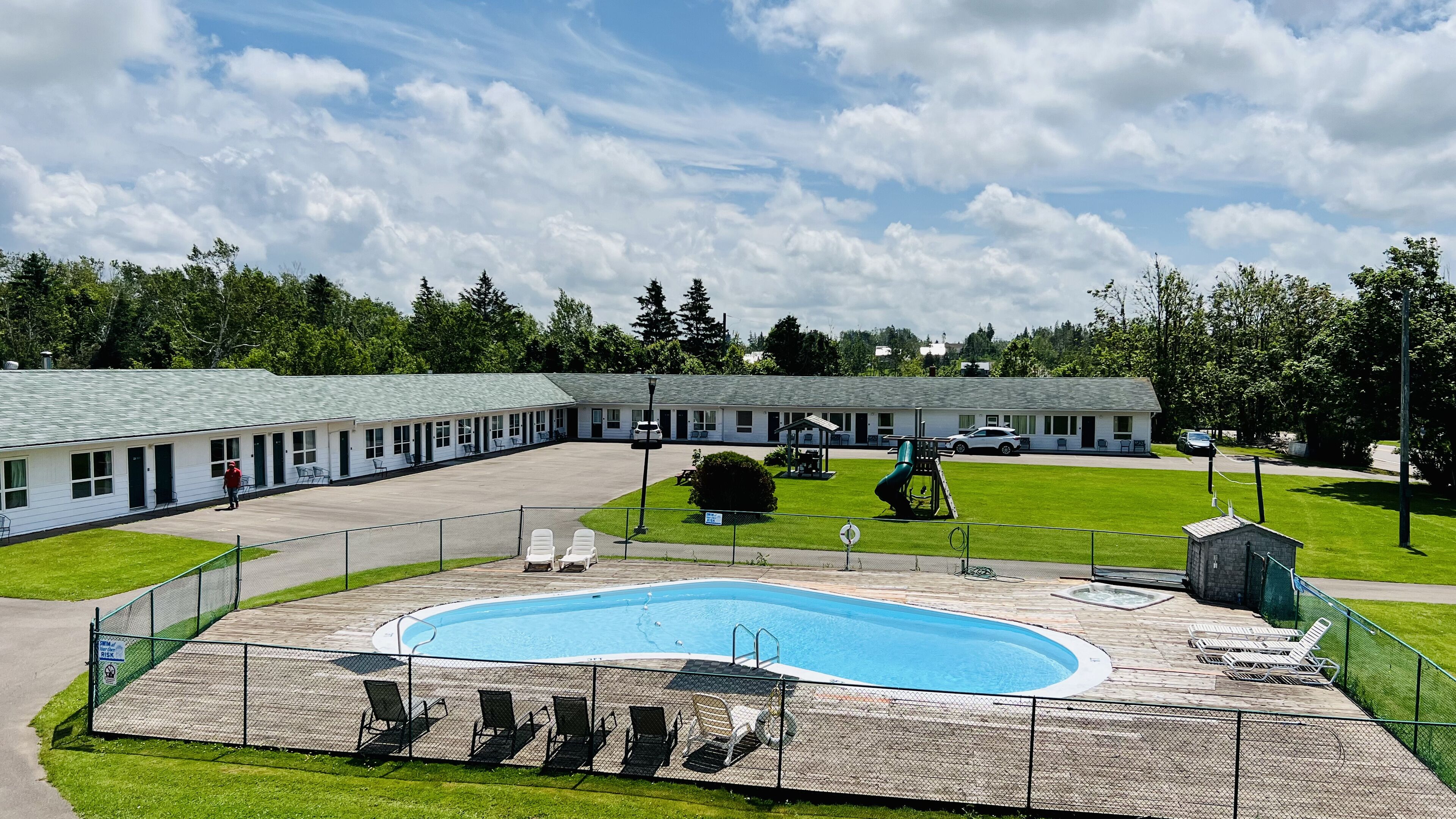 A motel with a swimming pool, a fenced deck area with lounge chairs, and a parking lot.