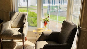 A room with two armchairs, a small table with a vase, and large windows offering a view of a residential area. - Anne Shirley Motel & Cottages (Cavendish)
