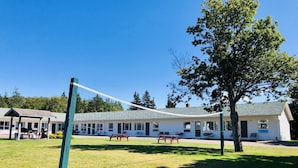 A sports court with a volleyball net, surrounded by a building with multiple windows and a covered seating area. - Anne Shirley Motel & Cottages (Cavendish)