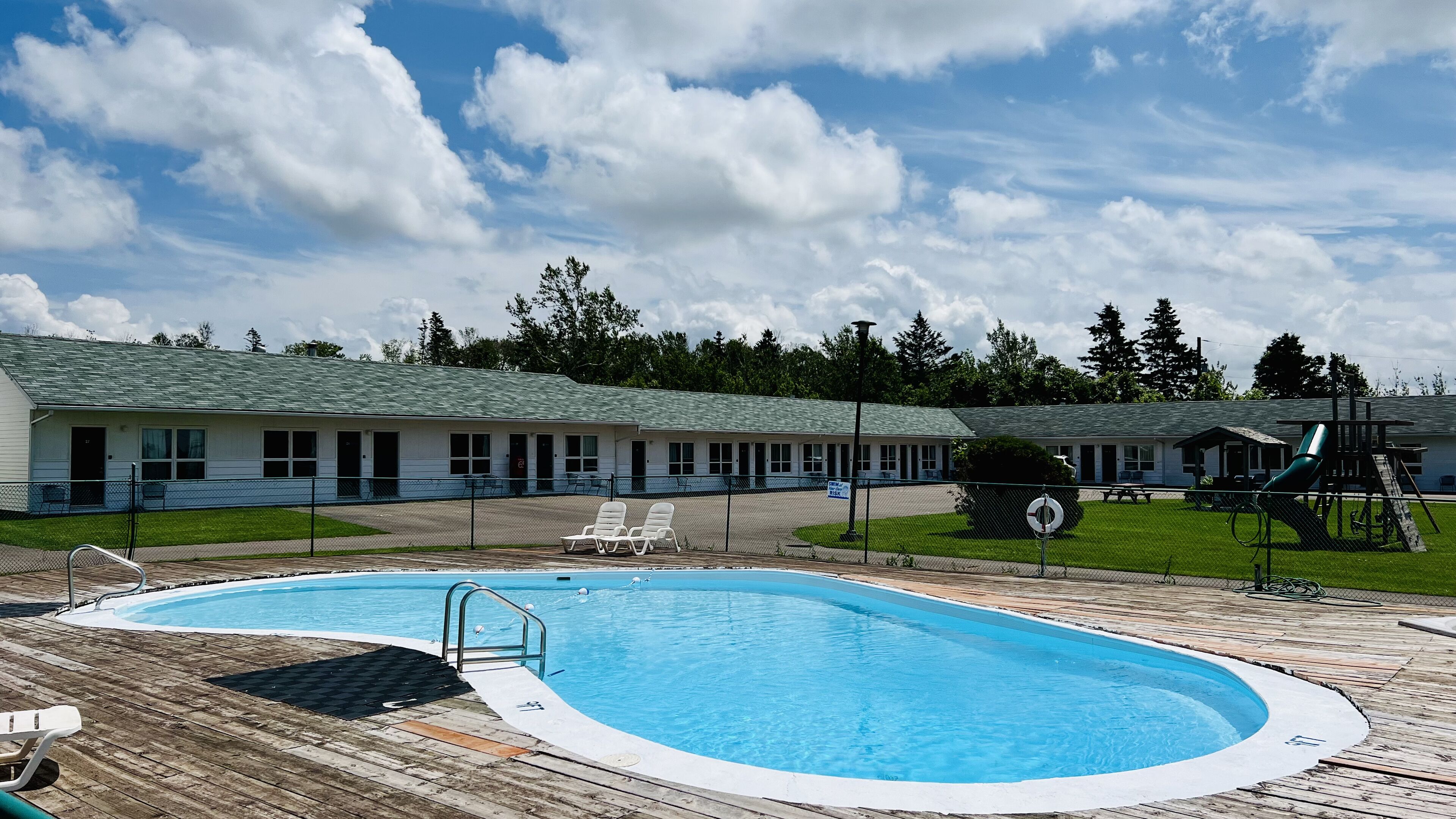 Una piscina con muelle de madera, sillas de descanso y un motel al fondo.