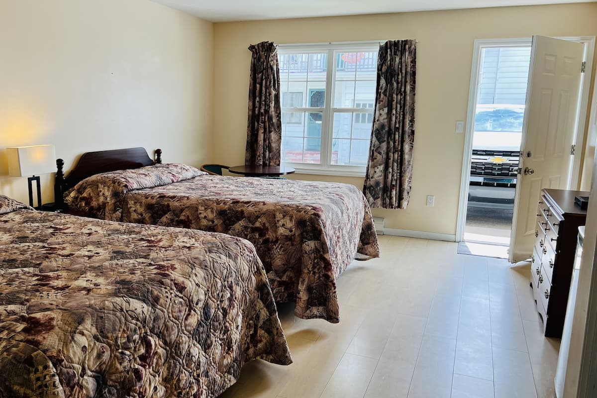 Una habitación con dos camas, una mesita, una silla, un aparador y una ventana con cortinas.