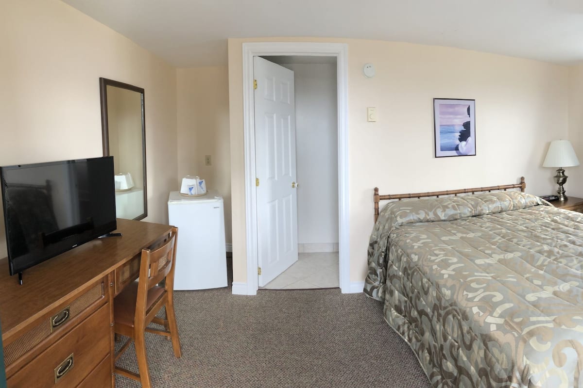 Una habitación de hotel con una cama, un escritorio con televisor, una mesita de noche, una lámpara y una ventana con cortinas.