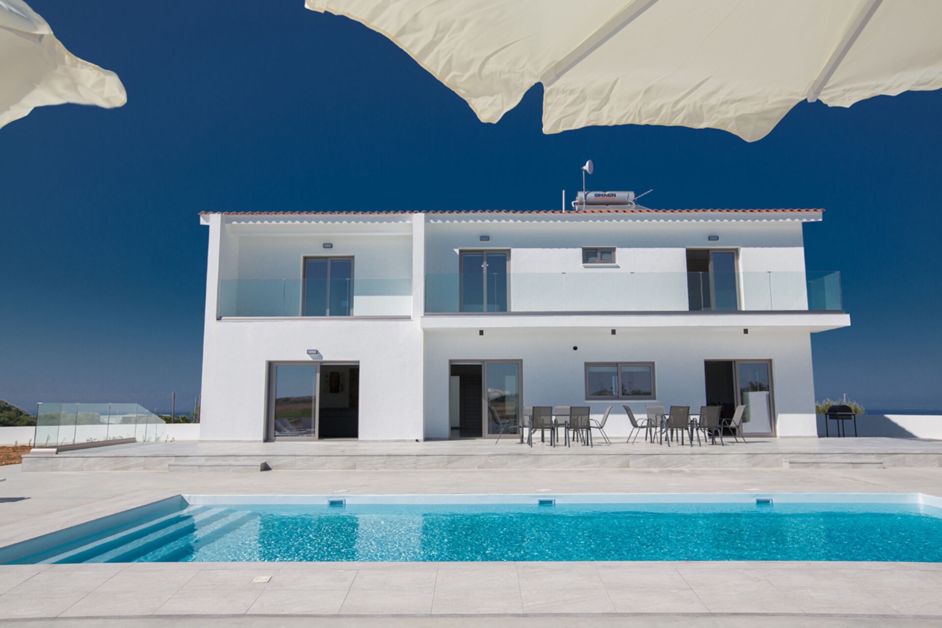 A two-story modern house with a swimming pool, outdoor furniture, and a clear blue sky.