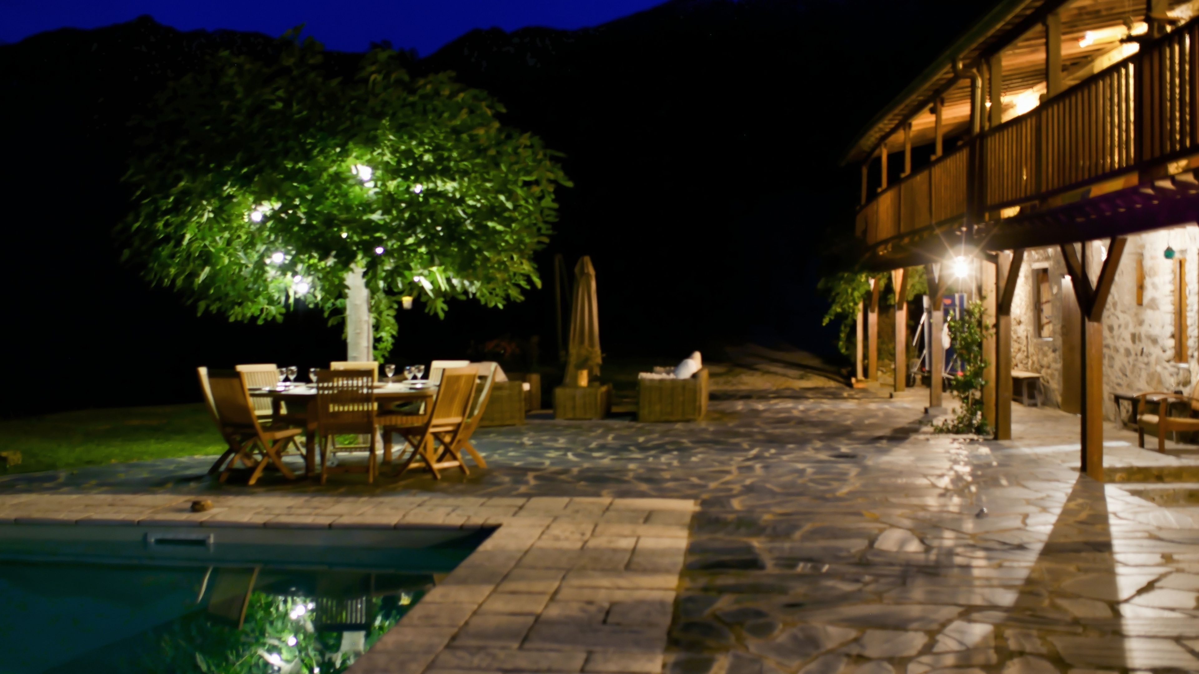 An outdoor dining area with a pool, stone flooring, and a tree providing shade.