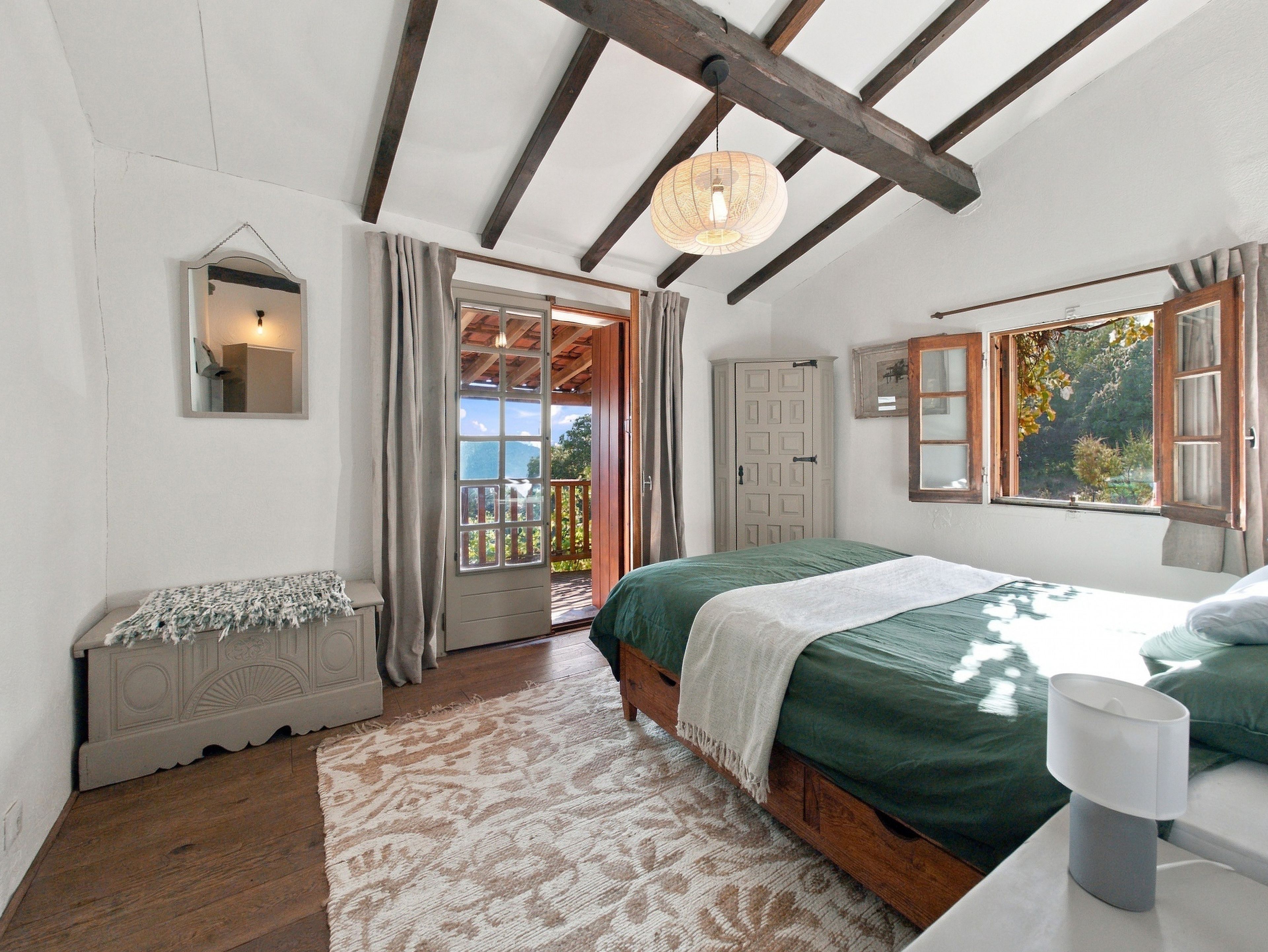 Une chambre à coucher comprenant un lit, une table de chevet, un miroir, une porte donnant sur un espace extérieur et une fenêtre avec vue sur la verdure.