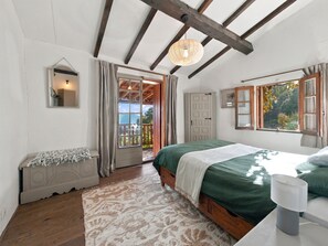 A bedroom with a bed, a nightstand, a mirror, a door leading to an outdoor area, and a window with a view of greenery.