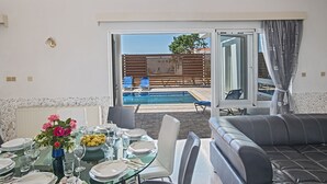 A dining area with a glass table set for a meal, a sofa, and a view of an outdoor swimming pool. - 5 Star Villa for Rent in Cyprus, Ayia Napa Villa 1201 (Ayia Napa)