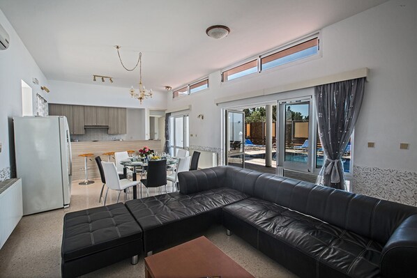 A modern living room with a black leather sofa, a dining area with a glass table, and a kitchen in the background.
