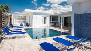 A pool area with blue lounge chairs, a clear sky, and a white building with a tiled wall. - 5 Star Villa for Rent in Cyprus, Ayia Napa Villa 1201 (Ayia Napa)