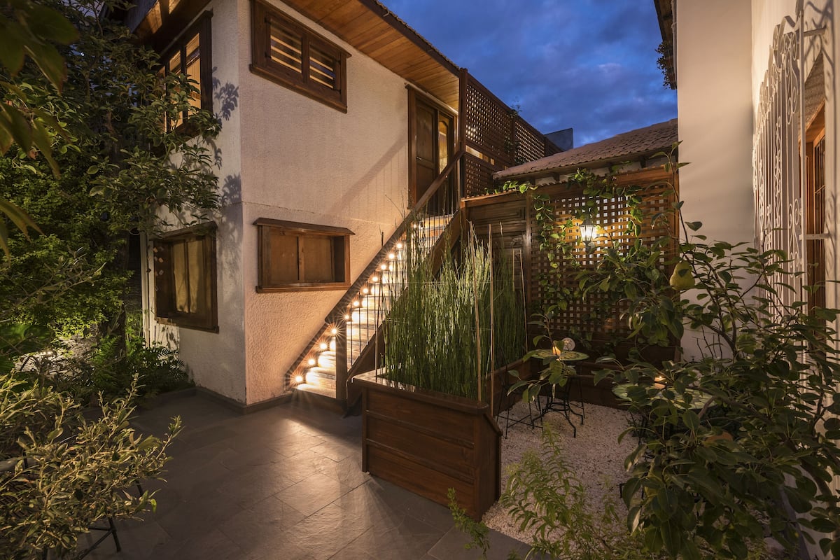 Un escalier bien éclairé mène à une terrasse en bois agrémentée d’une rambarde, le tout étant entouré de verdure et bordé d’un bâtiment blanc aux éléments décoratifs en bois.