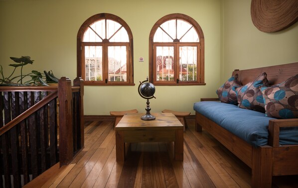 Una habitación con suelo de madera, una mesa de madera con un globo terráqueo, un sofá azul con cojines estampados y dos ventanas arqueadas con vistas a edificios.