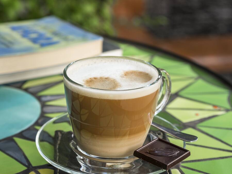 A glass cup of coffee with a chocolate piece on a coaster, placed on a green table with a book in the background.