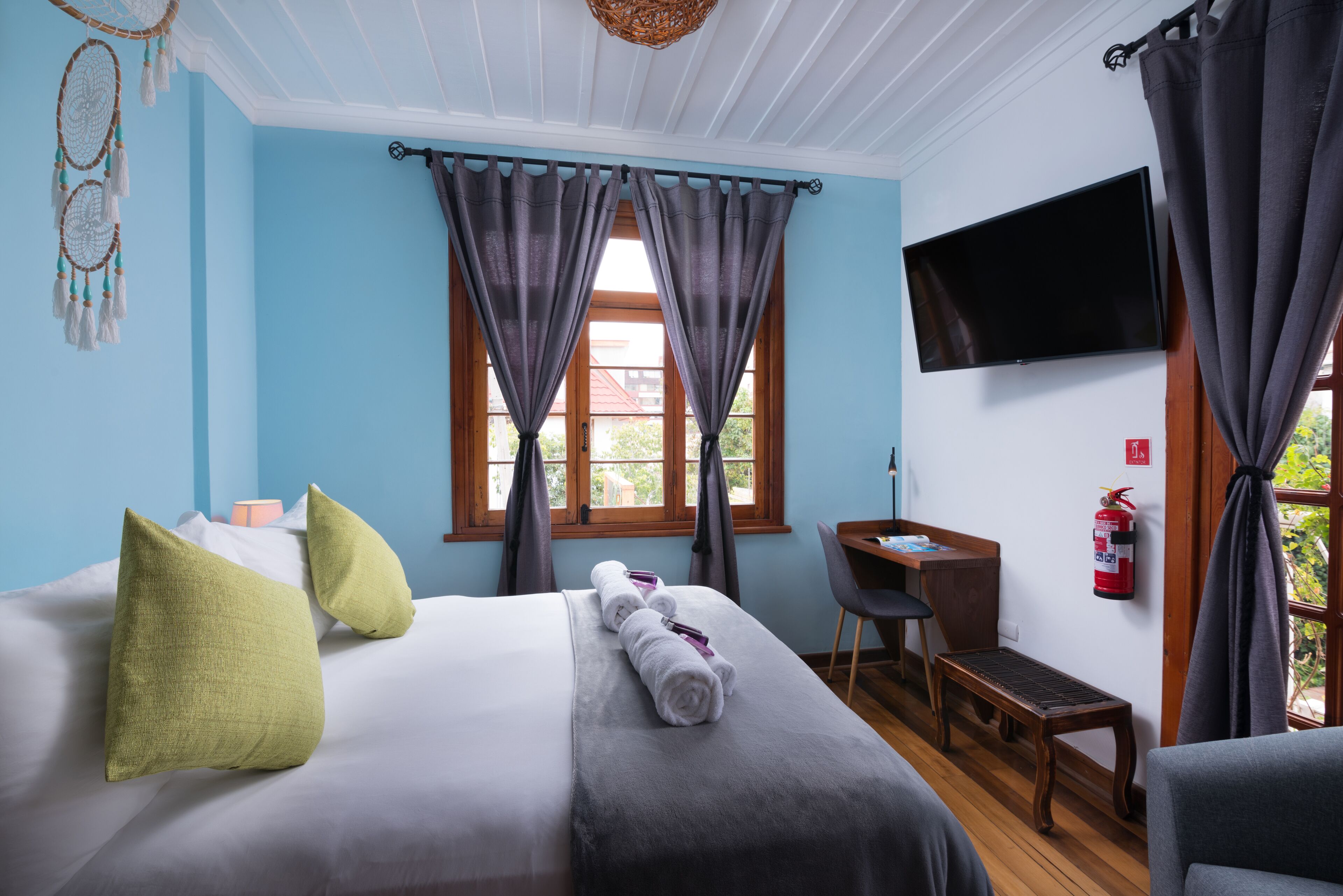 Quarto de hotel com cama, escrivaninha, cadeira, televisão e janela com cortinas.