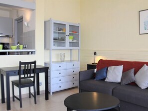 Un espacio habitable compacto con kitchenette, una zona de comedor con una silla y mesa, un sofá gris con cojines y una pequeña mesa redonda.