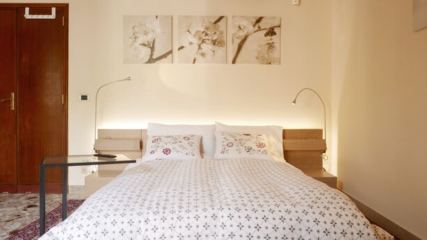 A bedroom with a bed, a nightstand, a bedside table, a lamp, and a door. - Three Rooms (Messina)