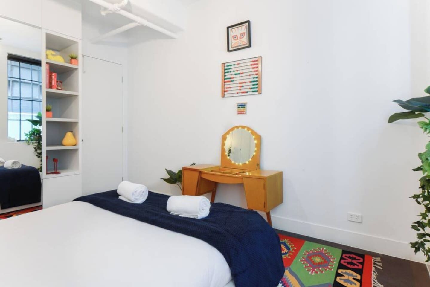 Un dormitorio con una cama, un aparador de madera con espejo, una estantería con libros y objetos decorativos, y una alfombra colorida.