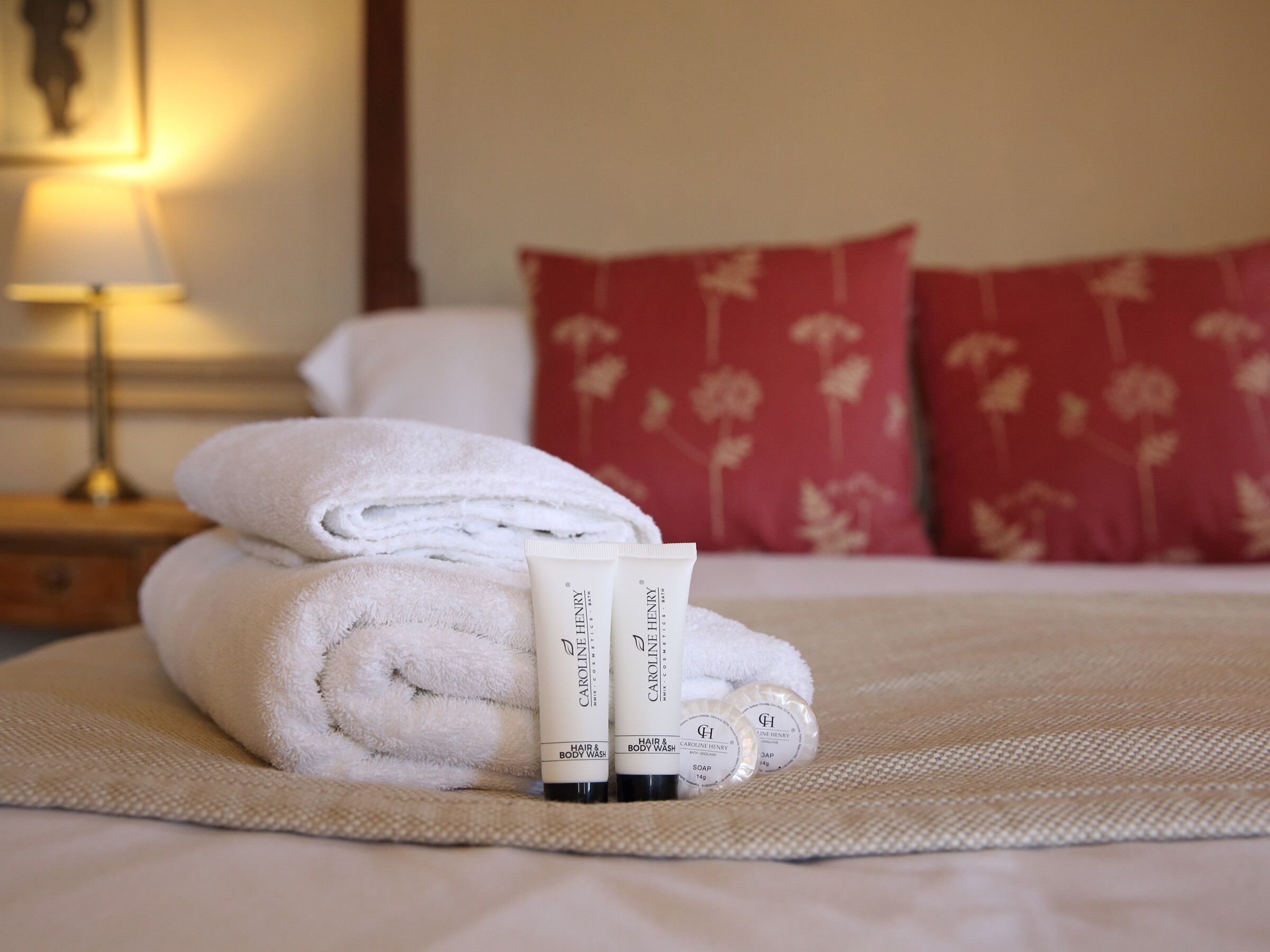 Una cama bien hecha con toallas blancas enrolladas, dos botellas de lavavásos y acondicionador para cabello y cuerpo Caroline Henry, y una almohada con estampado floral rojo.