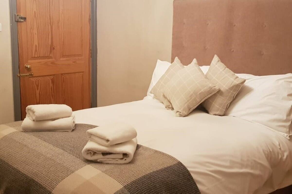 A neatly made bed with a checkered bedspread and folded towels, a wooden door, and a headboard with pillows.