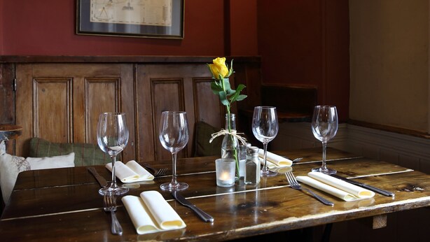 A dining table set with wine glasses, a vase with a yellow rose, salt and pepper shakers, and folded napkins. - Buckinghamshire Arms (Norwich)