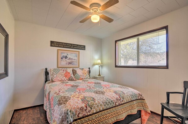 A bedroom with a bed, a ceiling fan, a window with blinds, a bedside lamp, and a framed picture on the wall.
