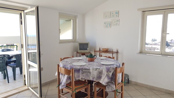 A dining area with a round table set for four, a television on a stand, and a window with a view of the city. - LUMINOUS SEA VIEWS IN SARDINIA, IDEAL FOR COUPLES OF FRIENDS AND FAMILIES! (Cala Gonone)