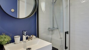 A bathroom with a white sink, a round mirror, a glass shower enclosure, and a blue accent wall. - The Alcove Royale, furnished with 3 stars tourism (Versailles)