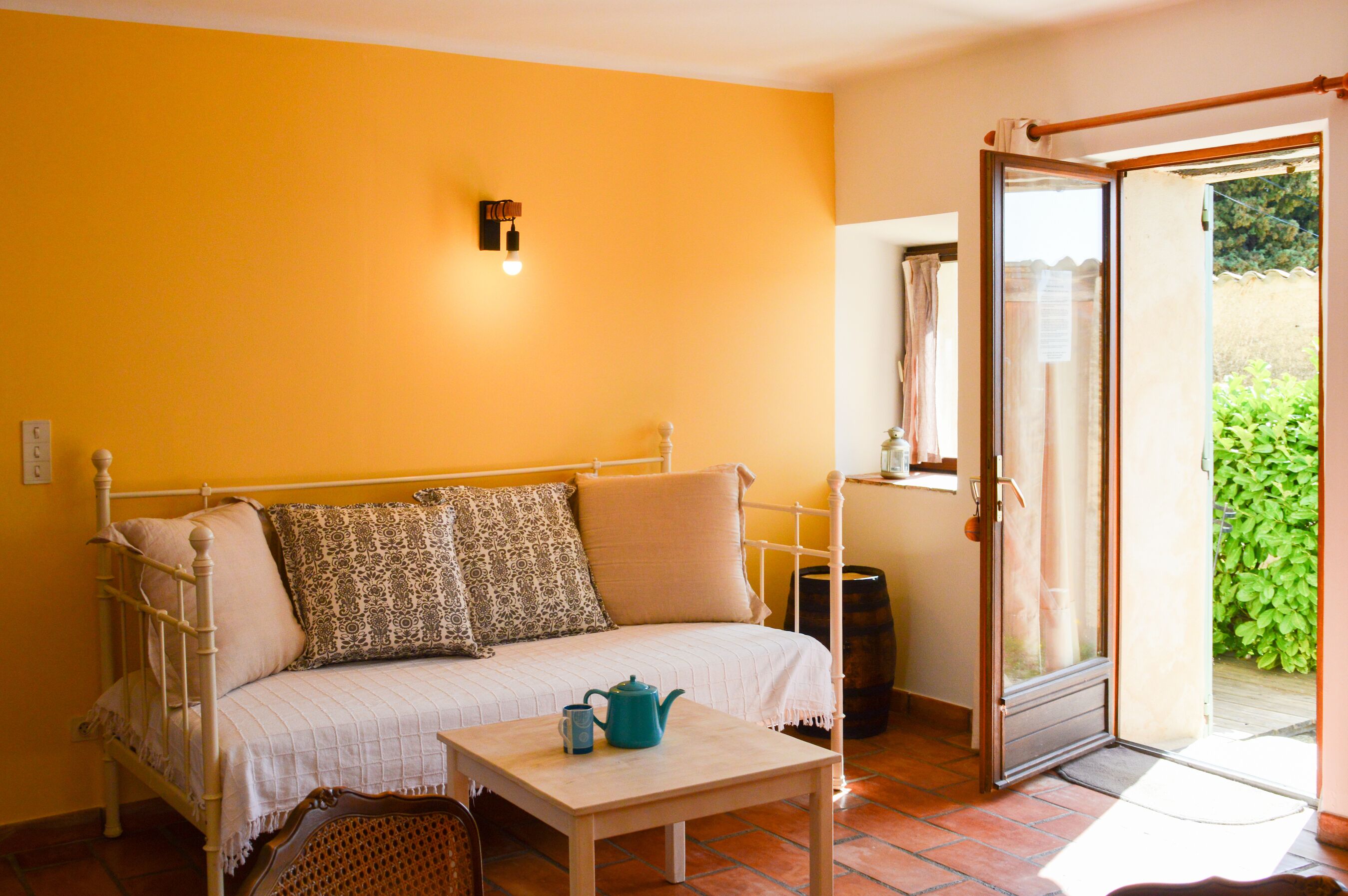 A room with a bed, a table with a teapot and cup, a door leading to an outdoor area, and a wall-mounted light fixture.