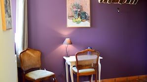 Une pièce aux murs violets, ornée d’un tableau représentant des fruits et des fleurs, avec deux chaises en bois, une table blanche et une lampe.