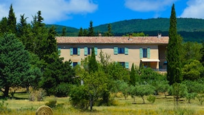 Un bâtiment à deux étages, doté d’un toit en tuiles, entouré d’arbres et d’un champ herbeux.